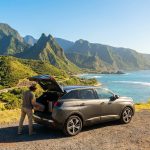 Un voyageur charge ses bagages dans une voiture de location face à un paysage côtier montagneux à la Réunion.