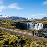 Un aventurier debout à côté d'un 4x4 vert devant une cascade islandaise au pied des montagnes enneigées.