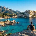 Un voyageur avec un sac à dos contemple Bonifacio et ses falaises surplombant la mer Méditerranée.