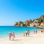 Famille et vacanciers marchant sur une plage de sable blanc au pied d'un village historique en Méditerranée.