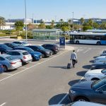 Un voyageur avec une valise marchant dans un parking d'aéroport avec de nombreuses voitures garées et un bus.