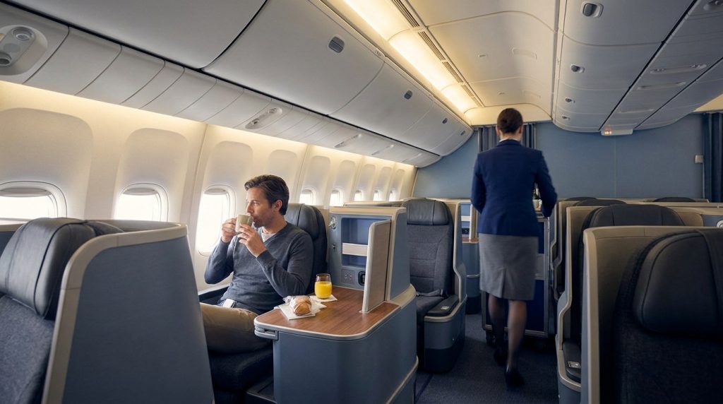 Passager assis en classe affaires dans un avion, buvant un café tandis qu'une hôtesse de l'air passe dans l'allée.