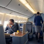 Passager assis en classe affaires dans un avion, buvant un café tandis qu'une hôtesse de l'air passe dans l'allée.