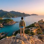 Un voyageur avec un sac à dos contemplant une côte rocheuse et une mer turquoise au coucher du soleil.