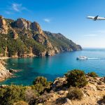 Un avion survole un ferry naviguant dans une baie aux eaux turquoise près des falaises escarpées de la Corse.