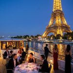 Couple et invités dînant sur le pont d'un bateau sur la Seine avec la Tour Eiffel illuminée en arrière-plan.