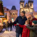 Une famille souriante consulte une carte sur un smartphone devant un marché de Noël à Prague.
