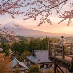 Une personne sur une terrasse en bois admirant le mont Fuji avec des cerisiers en fleurs au premier plan.