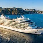 Un navire de croisière luxueux nommé Oceanic Elegance navigue sur une mer bleue près d'une côte montagneuse.