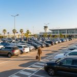 Un voyageur marche avec sa valise dans le parking extérieur ensoleillé de l'aéroport de Montpellier.
