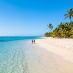 Un couple marche main dans la main sur une plage de sable blanc bordée de palmiers sous un ciel bleu à l'île Maurice.
