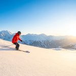 Un skieur en veste rouge dévale une pente enneigée face à un panorama de montagnes sous un soleil éclatant.