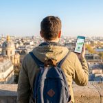 Un voyageur avec un sac à dos regarde Paris depuis une hauteur en tenant un smartphone affichant une application bancaire.