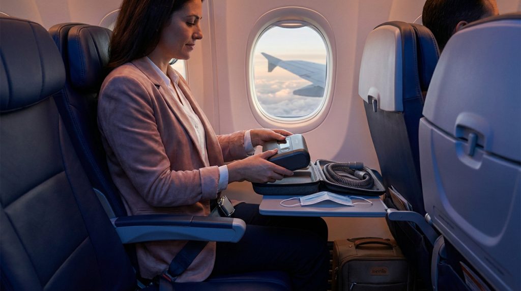 Une femme assise dans un avion tenant son appareil d'apnée du sommeil sur sa tablette devant une fenêtre.