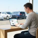 Un homme utilise un comparateur de location de voiture sur son ordinateur portable devant une vitrine de concessionnaire.