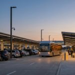 Des voyageurs avec des valises marchent sur un trottoir éclairé d'un parking d'aéroport avec des caméras de surveillance.
