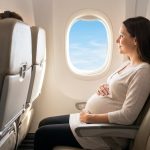 Une femme enceinte souriante assise près du hublot dans un avion, tenant délicatement son ventre.