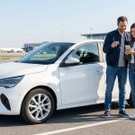 Un couple souriant récupère une voiture de location blanche sur le parking d'un aéroport.