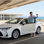 Un homme souriant debout près d'une voiture de location blanche avec les clés à la main sur un parking d'aéroport.