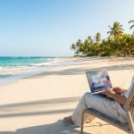Un homme utilise son ordinateur portable assis dans un fauteuil sur une plage paradisiaque au bord de l'eau.