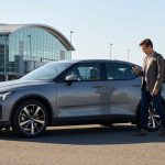 Un homme souriant consulte son téléphone près d'une voiture électrique garée devant l'aéroport de Lyon.