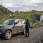 Un voyageur observe une cascade en Islande à côté d'un 4x4 Land Rover Defender garé sur un chemin de terre.
