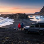 Deux voyageurs admirant le coucher de soleil sur une plage de sable noir en Islande à côté d'un 4x4.