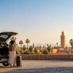 Une femme avec une valise devant sa voiture de location avec la mosquée Koutoubia de Marrakech en arrière-plan.