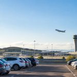 Voitures garées sur le parking de l'aéroport Lyon Saint-Exupéry avec un avion en plein décollage au-dessus de la tour.