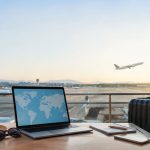 Ordinateur avec carte du monde, bagage et chapeau devant une piste d'aéroport au coucher du soleil.