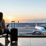 Une femme assise dans un aéroport devant une baie vitrée, regardant un avion au coucher du soleil avec sa valise.