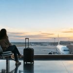 Une femme assise dans une salle d'embarquement d'aéroport avec une valise, regardant un avion au coucher du soleil.