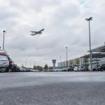 Vue au sol du parking P1 de l'aéroport avec des voitures garées et un avion en phase d'approche dans le ciel nuageux.