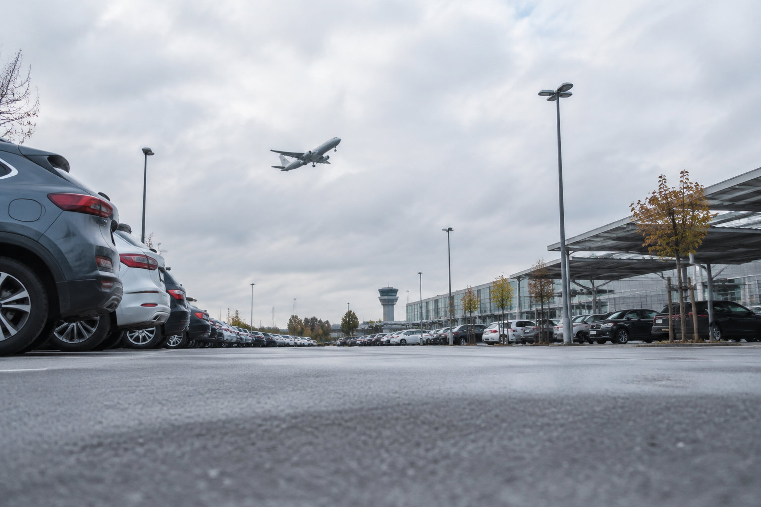 Vue au sol du parking P1 de l'aéroport avec des voitures garées et un avion en phase d'approche dans le ciel nuageux.