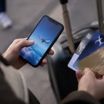 Une personne tient un smartphone et plusieurs cartes de fidélité aériennes devant une valise à roulettes.