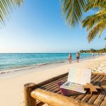 Passeport, chapeau et lunettes sur un transat avec une plage tropicale en arrière-plan.