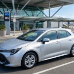 Une voiture hybride grise garée devant le terminal d'un aéroport avec une zone de récupération de location.